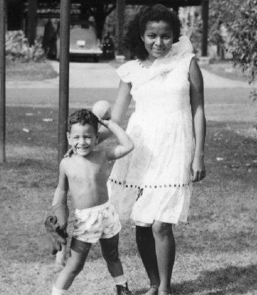 Vintage black-and-white photo of a smiling young boy holding a baseball and glove, standing beside a woman in a white dress, likely Dominga, on a lawn.