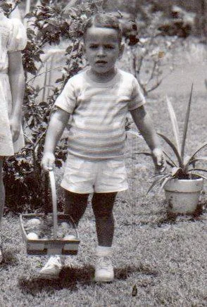 Vintage black-and-white photo of a small boy in a striped shirt and shorts carrying a basket across a garden lawn beside potted plants.