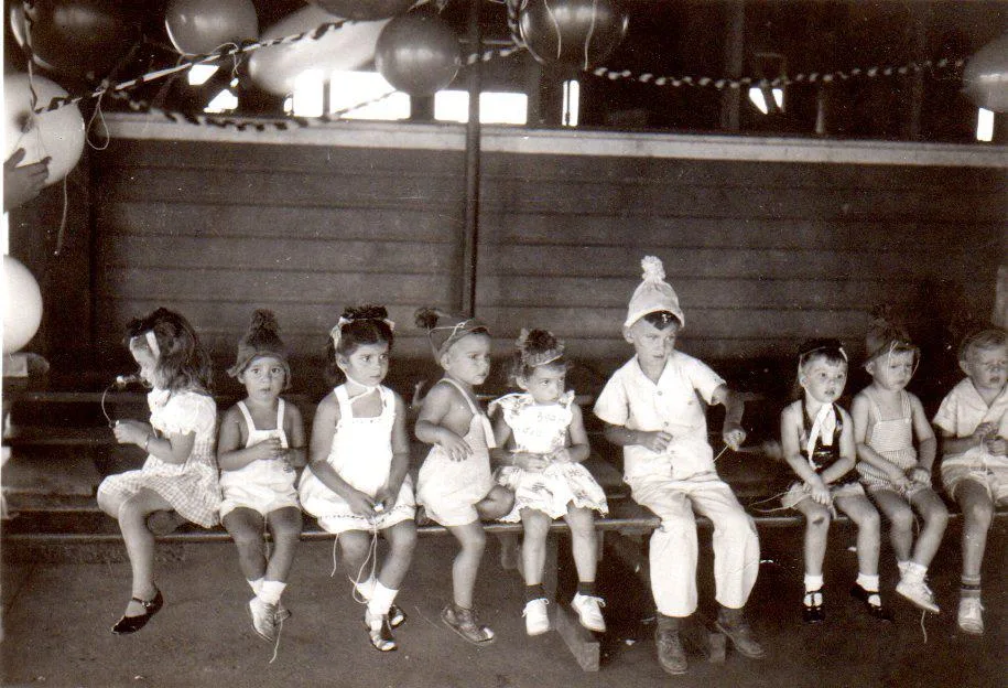 Vintage black-and-white photo of a row of young children in party hats seated on a bench beneath balloons and streamers.