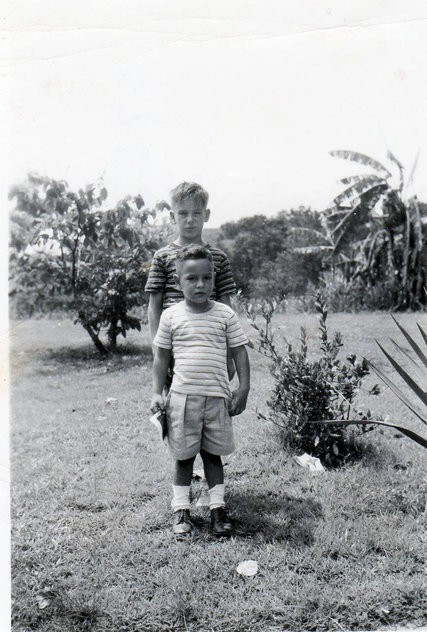 Vintage black-and-white photo of a small boy in a striped shirt standing in front of an older boy on grass, with banana plants and trees behind.