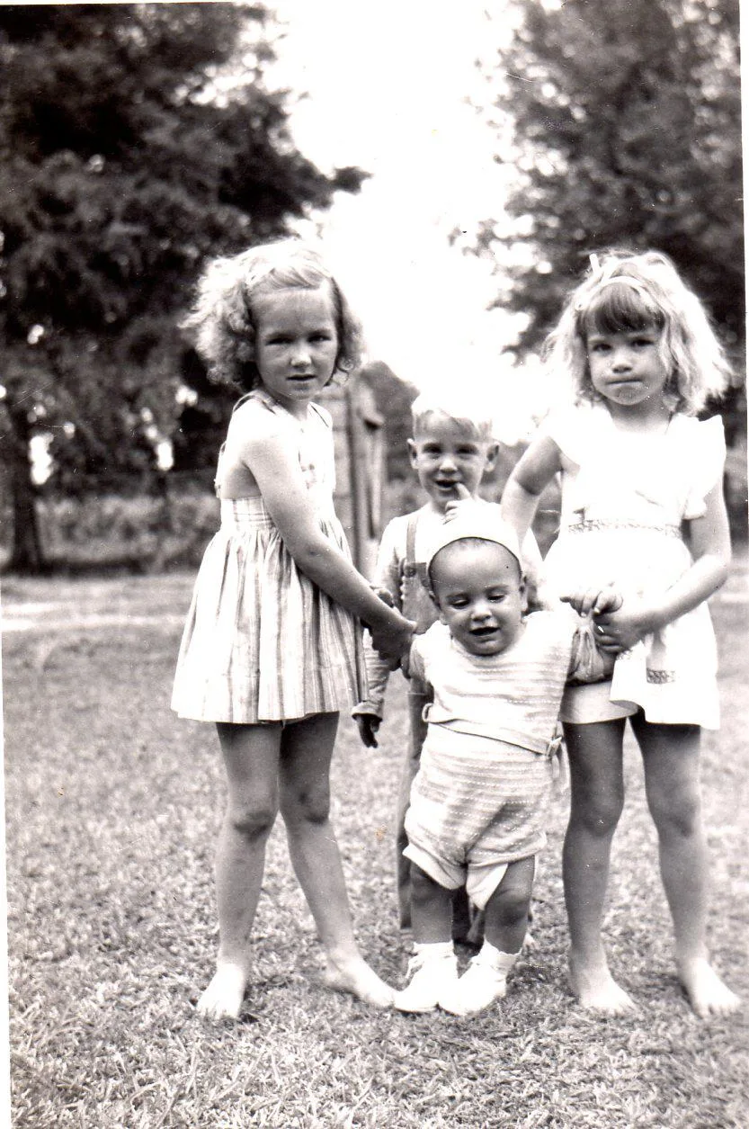 Vintage black-and-white photo of two young girls holding the hands of a smiling baby on grass, with a small boy standing behind them.
