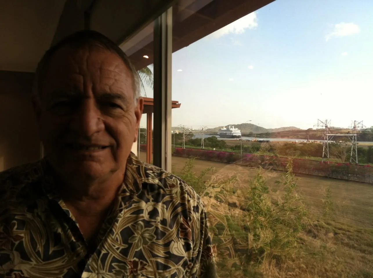 A smiling older man in a floral shirt stands by a large window overlooking the Panama Canal, with a cruise ship and green hills beyond.