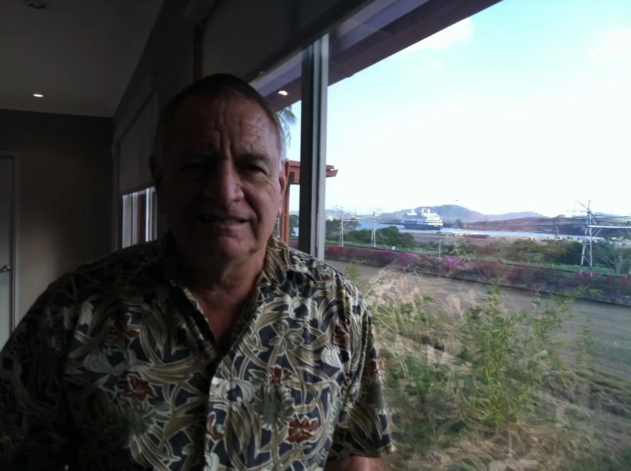 An older man in a floral shirt smiles indoors beside a window, with the Panama Canal and a cruise ship visible in the distance.