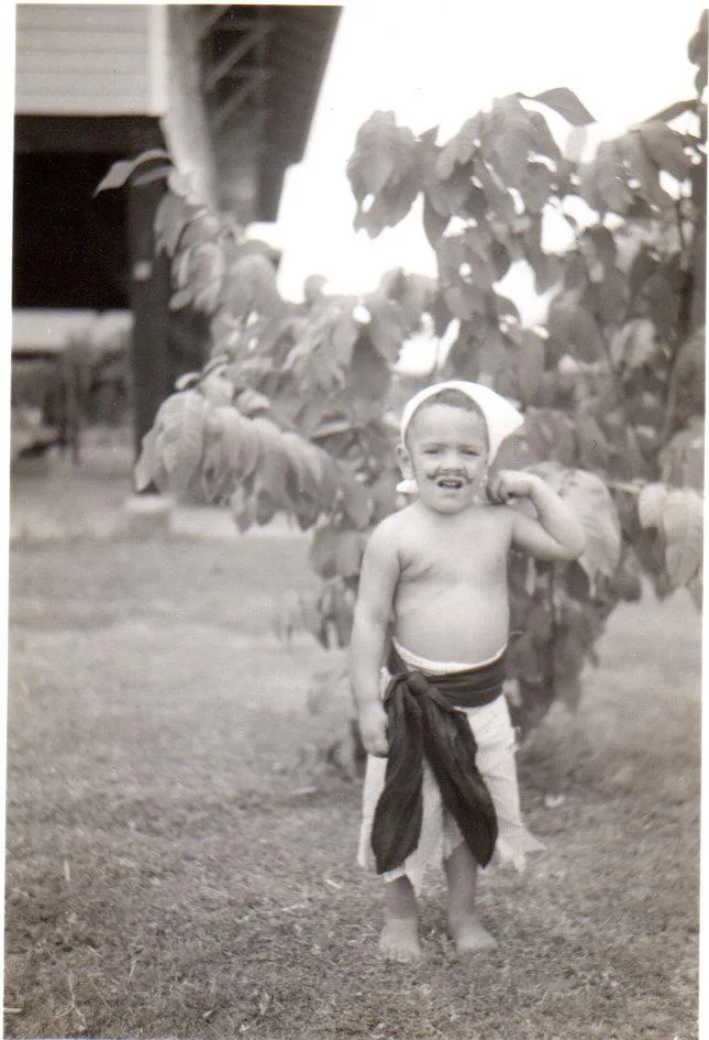 Black-and-white photo of a toddler in a white headscarf and sash skirt, flexing his arm and grinning on a lawn beside a leafy tree.