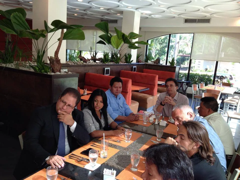 A group of men and a young woman seated at a long table in a bright restaurant with tropical plants, listening attentively during a lunch meeting.