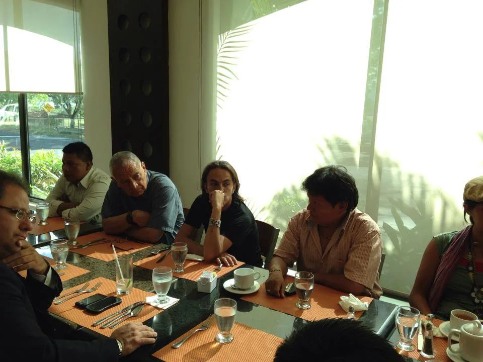 Participants at a lunch table by sunlit windows, among them an older man in a grey shirt and a man with long hair resting his chin on his hand.