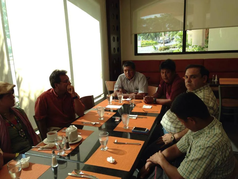 Indigenous leaders and others seated around a dining table with orange placemats and water glasses, one man speaking while others listen.