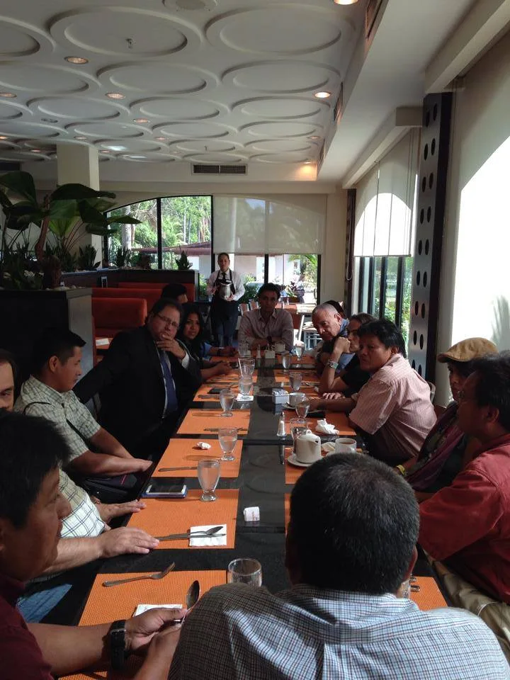 Long view of a crowded table in a restaurant with a patterned ceiling, many participants gathered in discussion as a server stands in the background.
