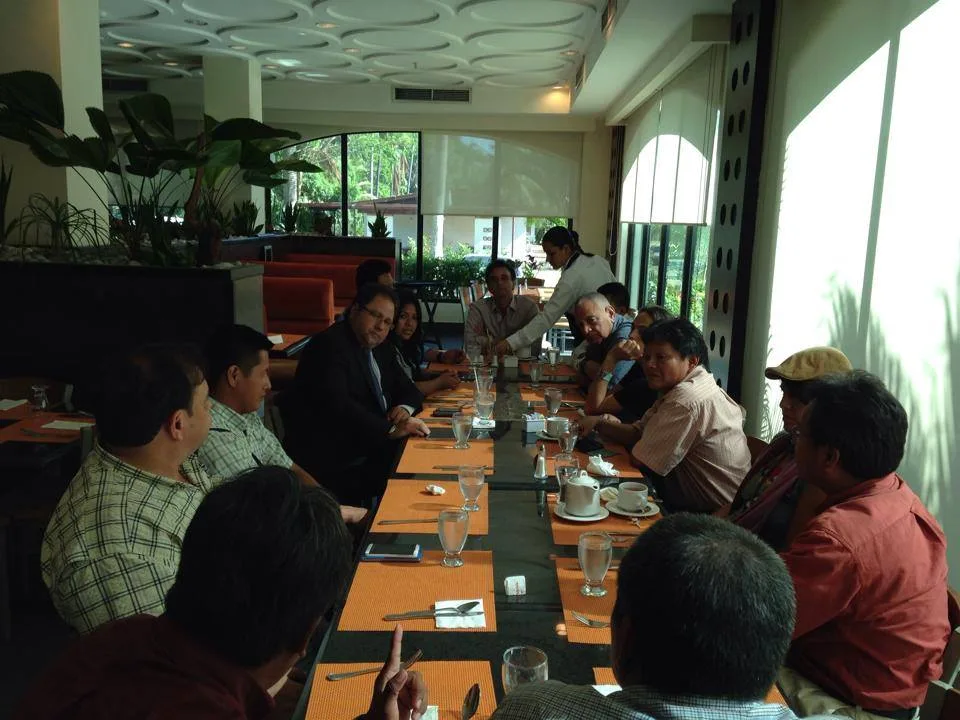 Wide view of the lunch meeting, with people seated along both sides of a long table while a server pours drinks at the far end.