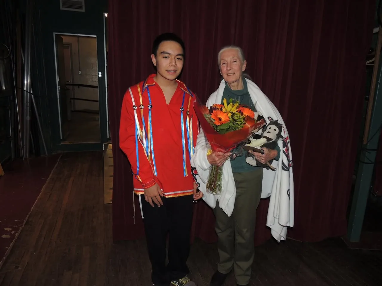 A young man in a red ribbon shirt stands beside Jane Goodall, who holds an orange bouquet and a stuffed monkey, with a white blanket over her shoulders, before a red stage curtain.