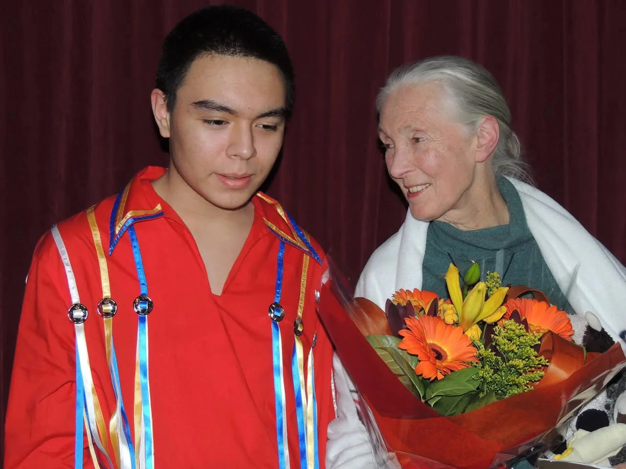 Jane Goodall smiles at Tiger, a young man in a red ribbon shirt, as she holds a bouquet of orange gerberas and yellow lilies in front of a red curtain.