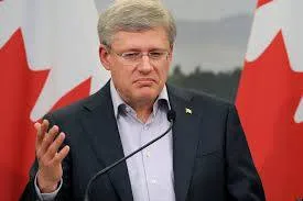 Prime Minister Stephen Harper in a dark suit standing at a microphone, gesturing with one hand, with Canadian flags behind him.