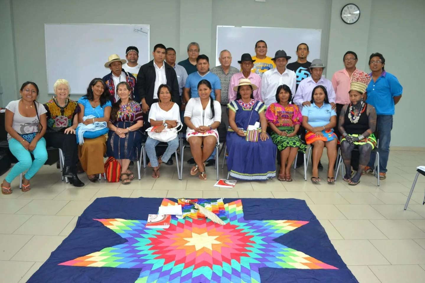 A large group of Indigenous men and women, some in traditional dress, pose in a classroom around a rainbow star quilt on the floor holding a feather fan and papers.