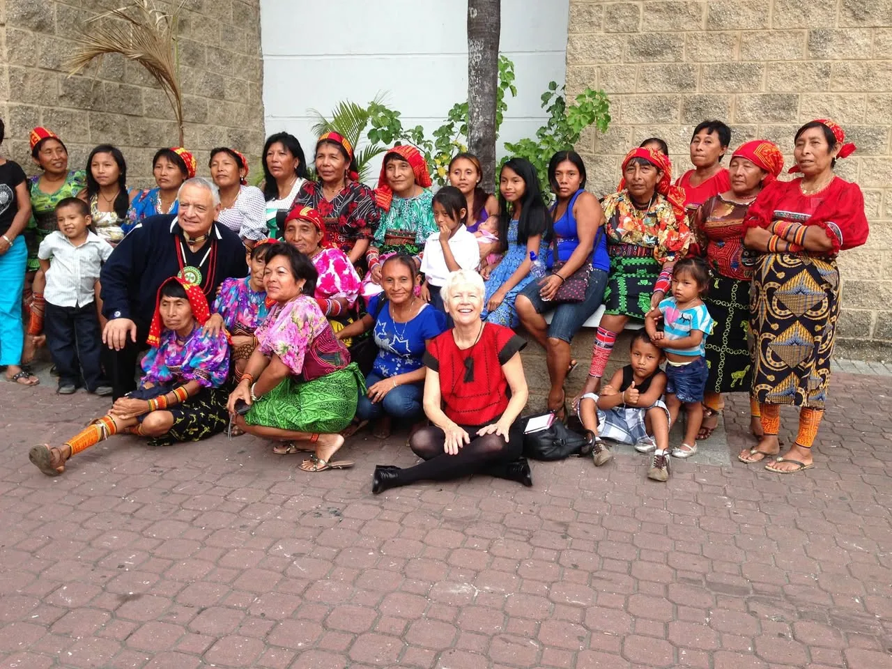 A large group of Guna women in red headscarves and colorful mola blouses, with children, pose outdoors on a brick plaza with an older man and a white-haired woman.