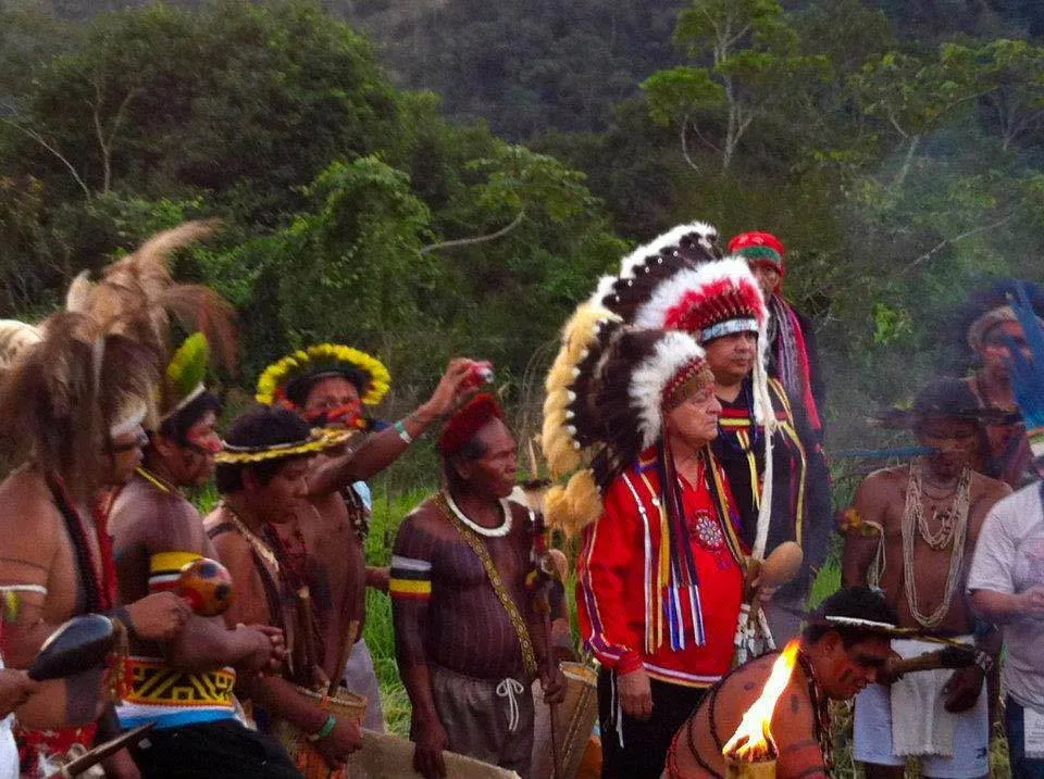 Indigenous men in feather headdresses and body paint gather outdoors near a forested hill, among them an elder in a red shirt and a large eagle-feather war bonnet.