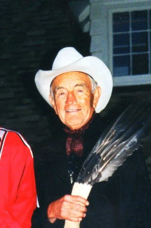 Phil Lane Sr., an elderly man in a white cowboy hat and dark scarf, smiling and holding an eagle feather fan outdoors at night.
