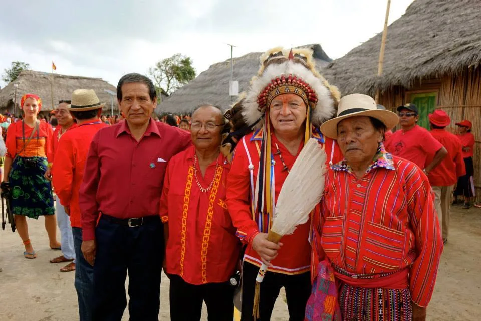 Phil Lane Jr. in a feathered headdress, holding a feather fan, stands with three Indigenous men in red shirts before thatched-roof houses.