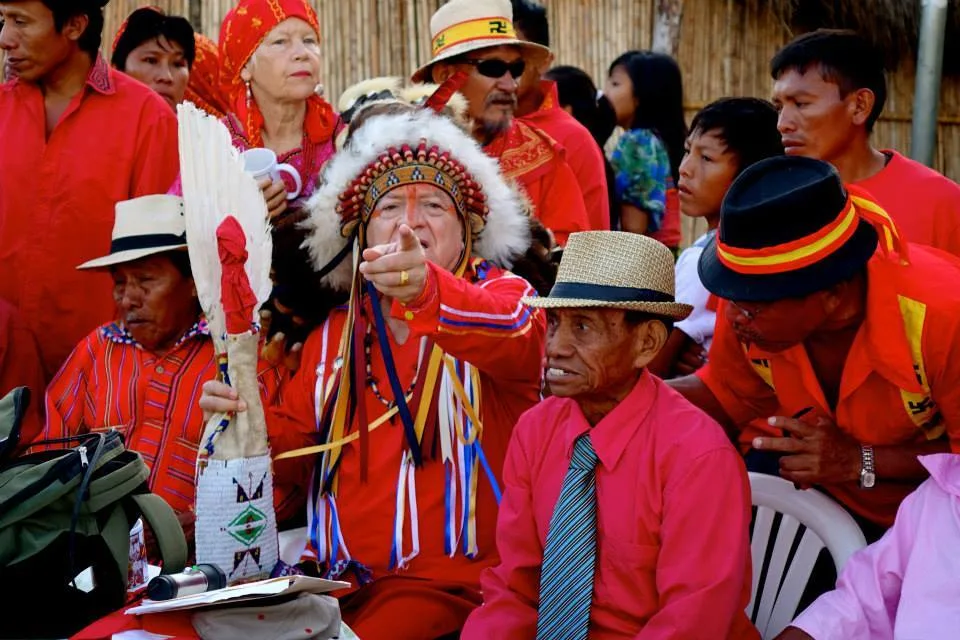 Phil Lane Jr. in a feathered headdress points toward something while seated among elders and community members dressed in red.
