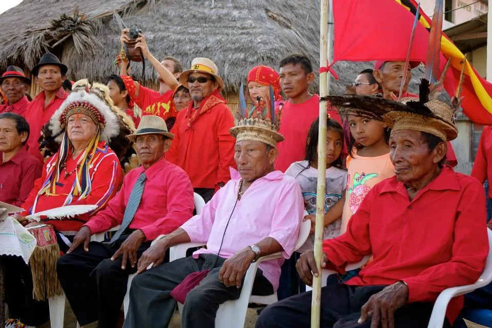 Phil Lane Jr. sits beside Guna elders, some in woven feathered hats, with a crowd in red clothing and a red flag behind them.