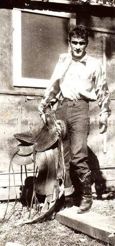 Vintage black-and-white photo of a young man in jeans and boots holding a saddle outside a wooden building.