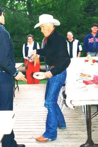 An elder in a white cowboy hat and jeans holds a plate at an outdoor gathering on a wooden deck as others look on.