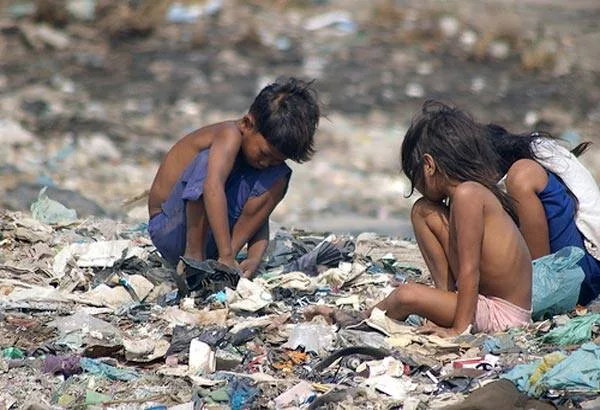 Young children crouching barefoot among garbage at a dump, sorting through the trash.
