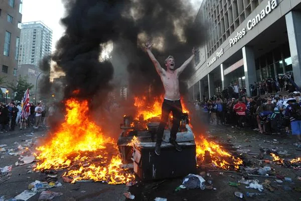 A shirtless man stands with arms raised atop a burning dumpster amid flames and black smoke on a littered city street, crowds watching beside a Canada Post building.