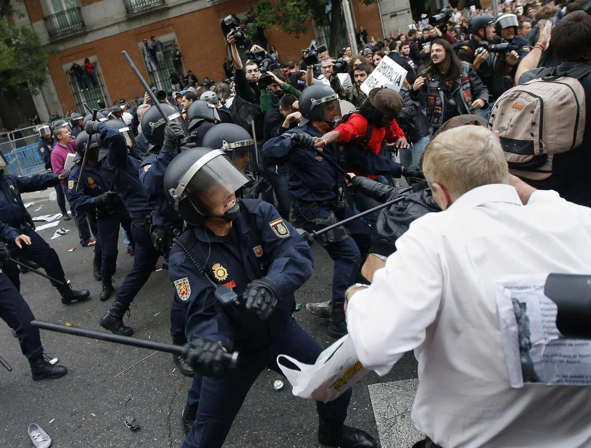 Riot police in helmets and visors swing batons at protesters in a crowded street as photographers film; a protester holds a sign reading "Democracia Real."