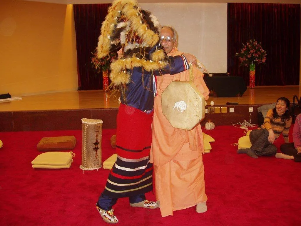 A dancer in Indigenous regalia with a fur and feather headdress and ribbon skirt embraces a smiling swami in saffron robes holding a hand drum, as seated people watch on a red carpet.