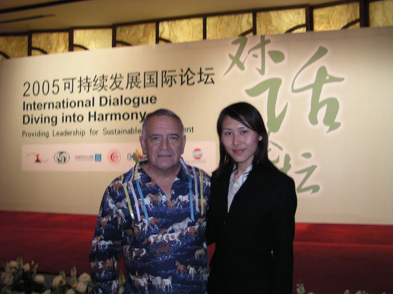 Phil Lane Jr., in a horse-print ribbon shirt, stands beside a young woman in a black suit before a banner reading 2005 International Dialogue, Diving into Harmony.