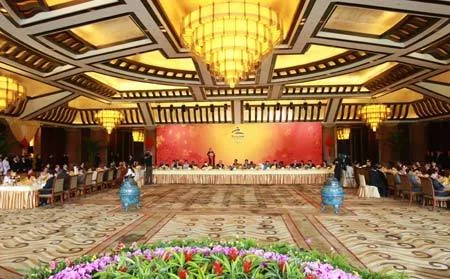 A grand banquet hall with chandeliers and an ornate ceiling, where delegates sit at long tables before a red backdrop.