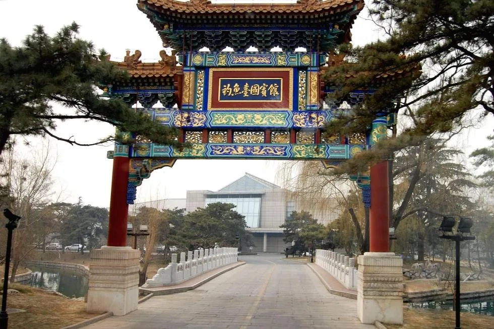 An ornate painted Chinese archway with red pillars and a gold inscription stands at the entrance to a bridge leading toward a modern building among pine trees.