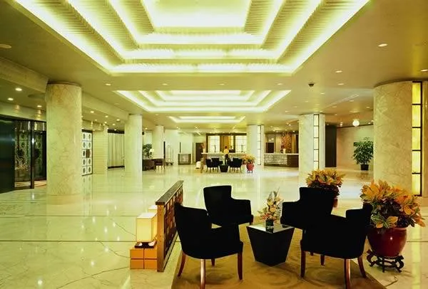 A spacious hotel lobby with marble columns and floors, glowing ceiling lights, black armchairs and potted plants.