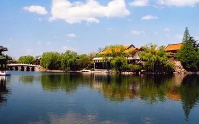 A calm lake reflecting willow trees and traditional buildings with golden roofs, with a stone bridge at left under a blue sky.