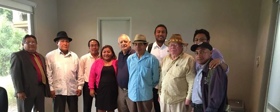 A group of ten men and women, several in hats and button-up shirts, stand together for a photo in an office room beside a window.