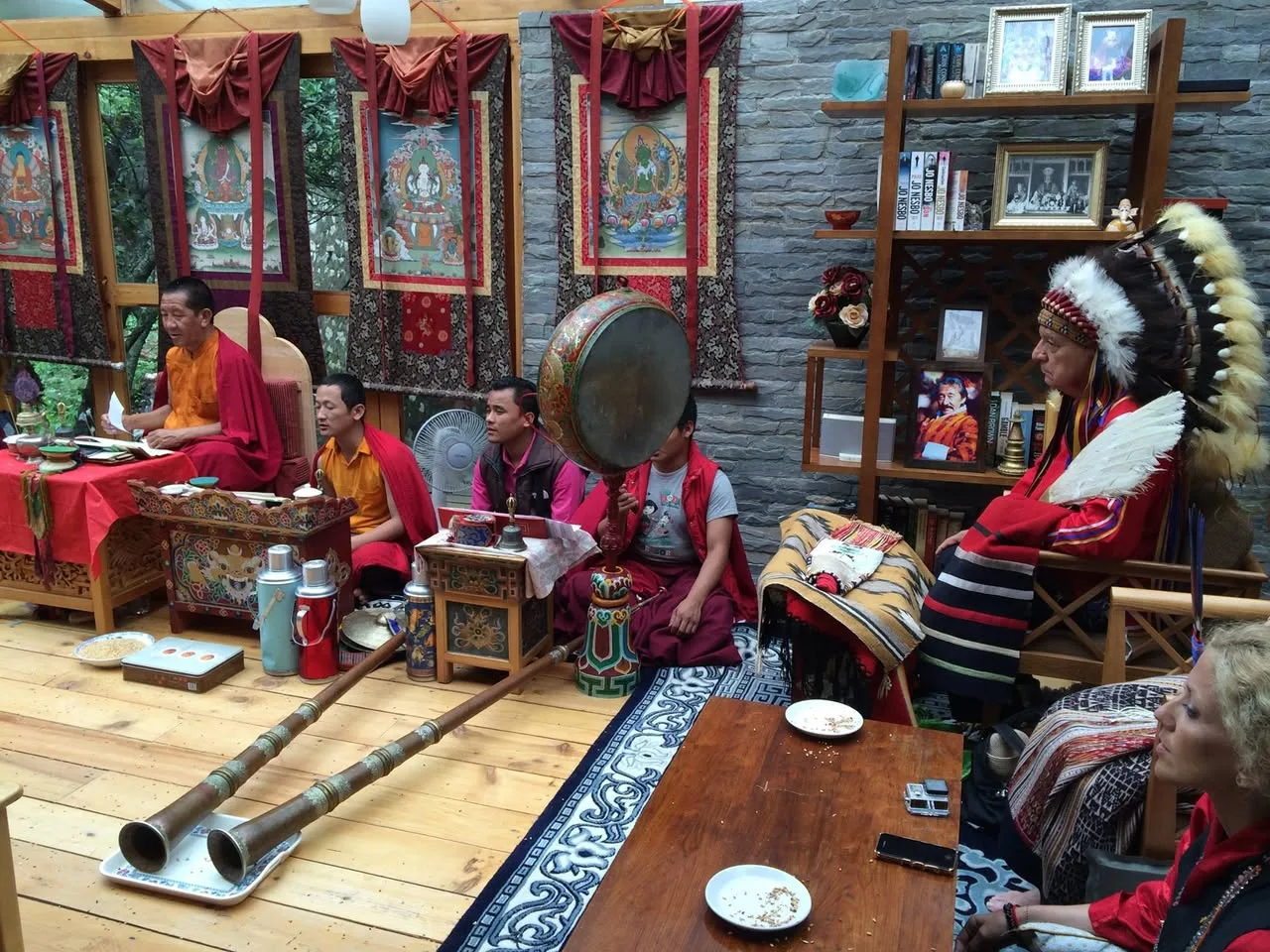 Buddhist monks in maroon robes chant and play a large drum beside long horns in a shrine room hung with thangkas, as an elder in a feather headdress sits watching.