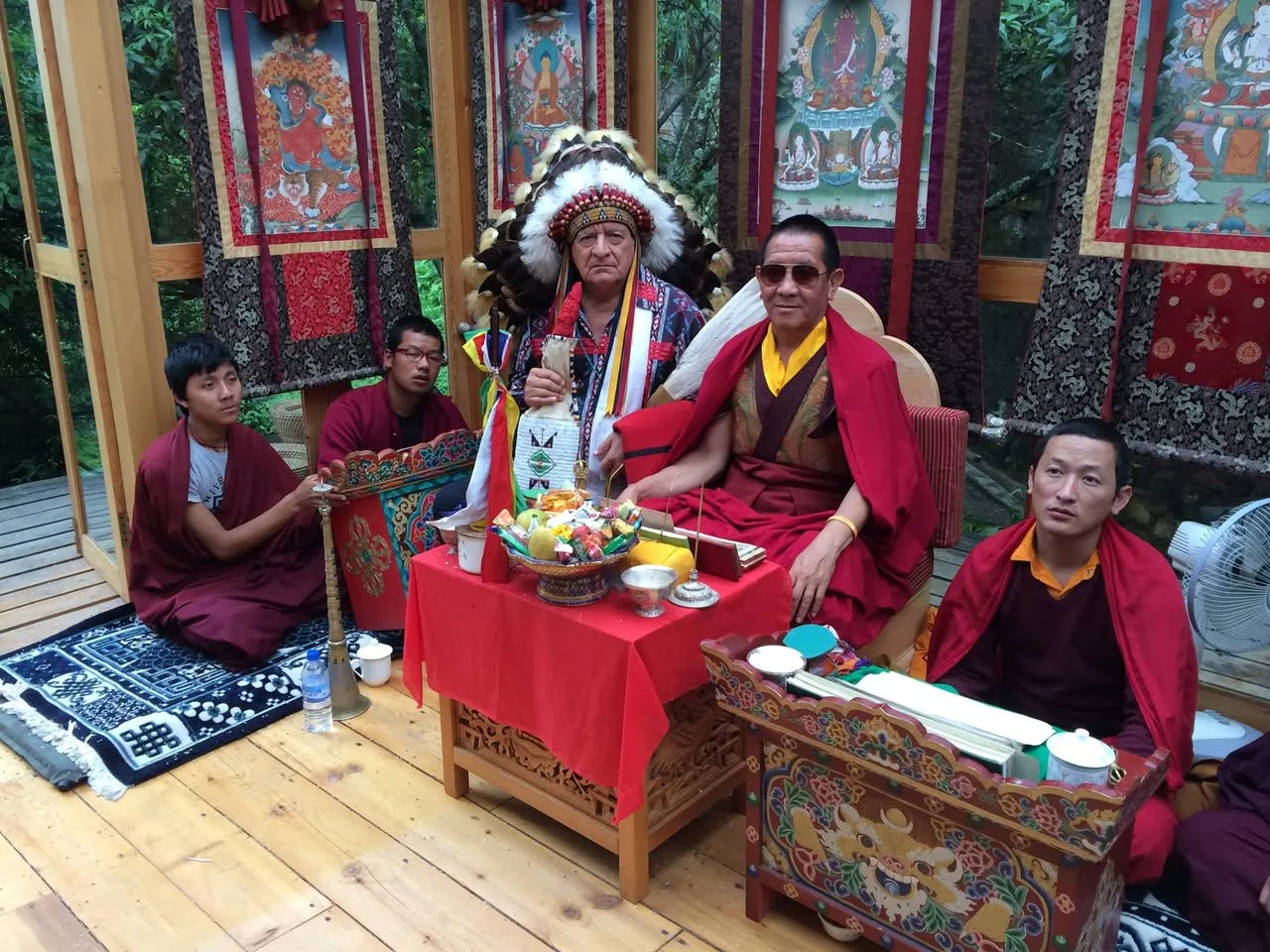 An elder in a full feather headdress holding a beaded bag sits beside a senior Buddhist lama in sunglasses, with young monks around an altar of offerings.