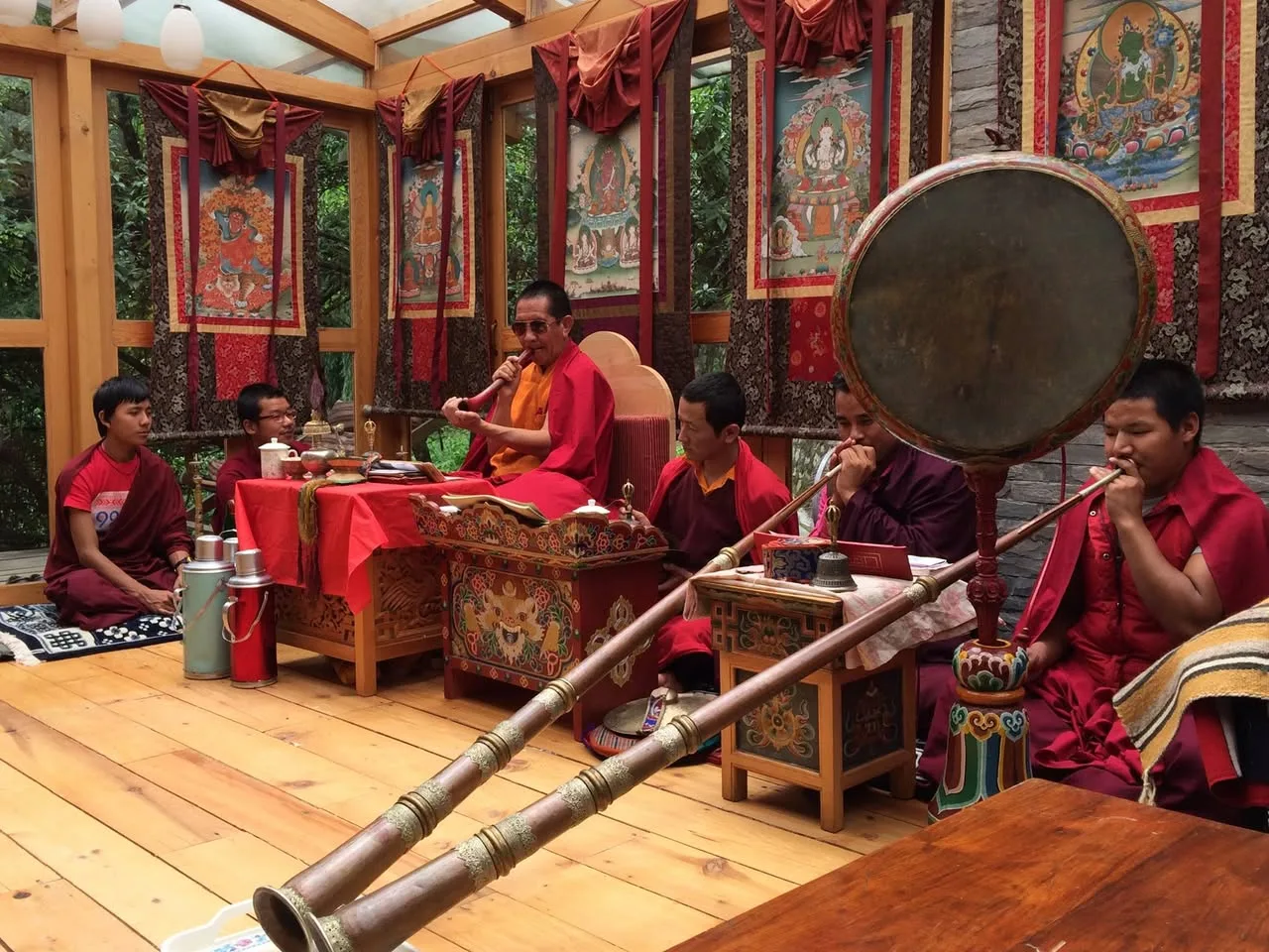 Buddhist monks in maroon and red robes play long horns and a large painted drum during a ceremony in a wooden shrine room hung with thangka paintings.