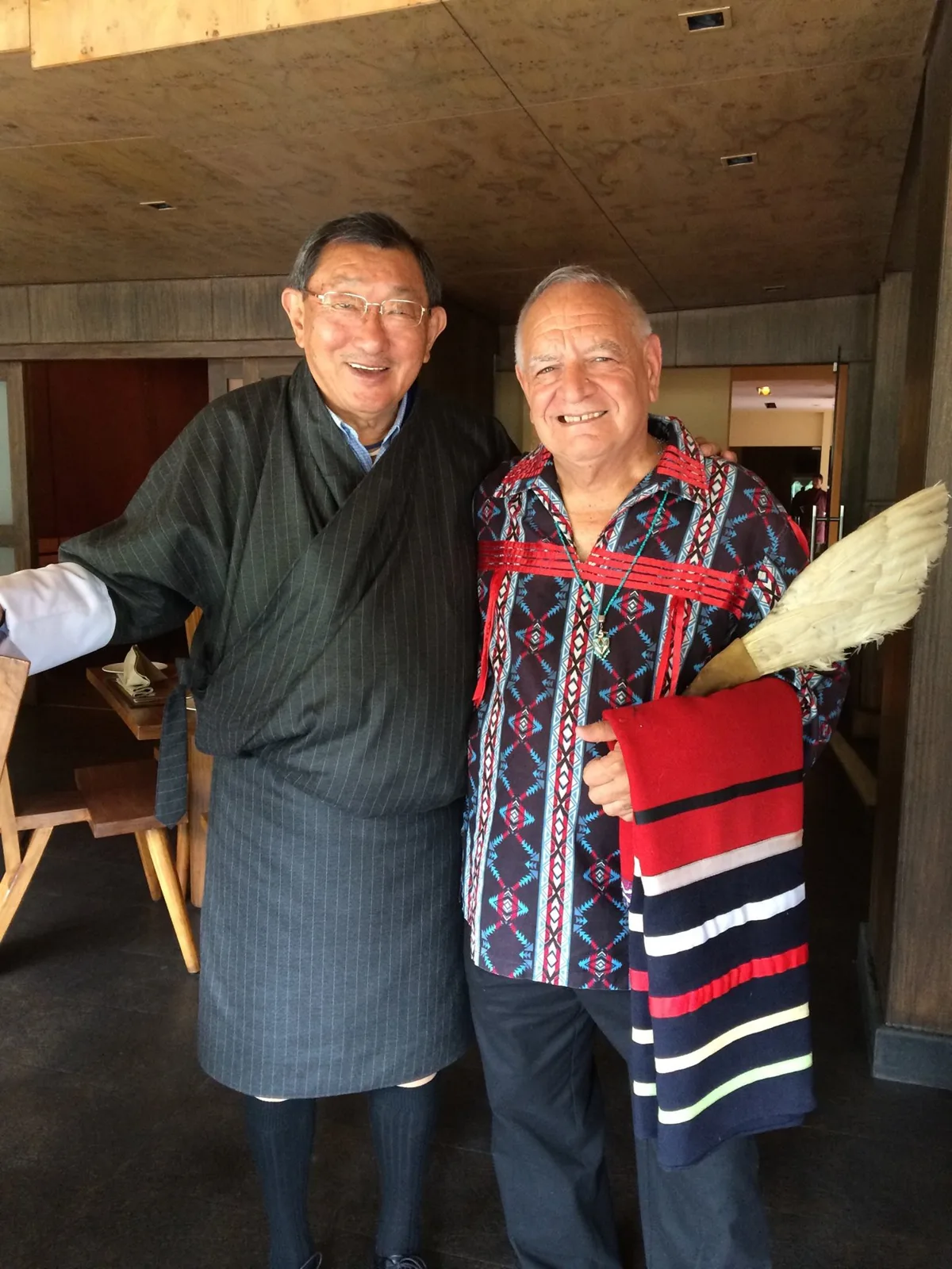 Two smiling elders stand arm in arm indoors, one in a dark striped Bhutanese robe, the other in a patterned ribbon shirt holding a striped blanket and a feather fan.