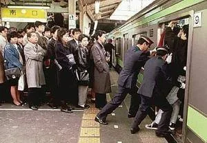 Uniformed station attendants push passengers into a packed commuter train while others wait in line on the platform.