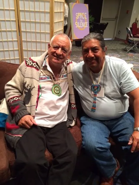 Two smiling Indigenous men sit side by side on a couch, one in a patterned cardigan with a green medallion, the other wearing a beaded medallion; a "Space for Silence" sign is behind them.