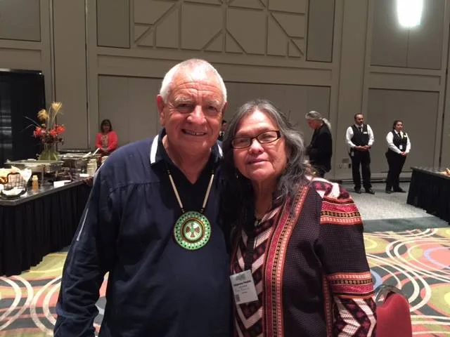 Phil Lane Jr., wearing a beaded medallion, stands smiling beside his sister Mona, in a patterned jacket and glasses, in a reception hall with buffet tables and servers behind them.