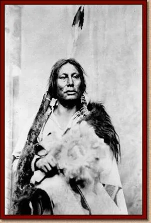 Historic black-and-white portrait of a Lakota man with long hair and a feather, wrapped in a fur robe and holding a feathered object.