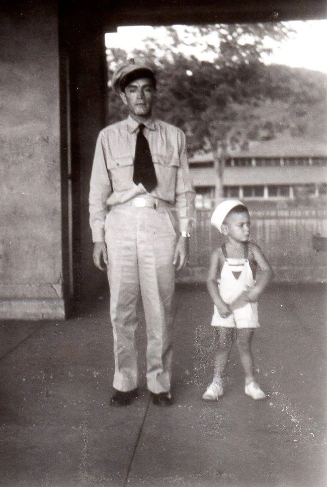 Vintage black-and-white photo of a man in a military uniform and cap standing beside a small boy in a white sailor hat and overalls.