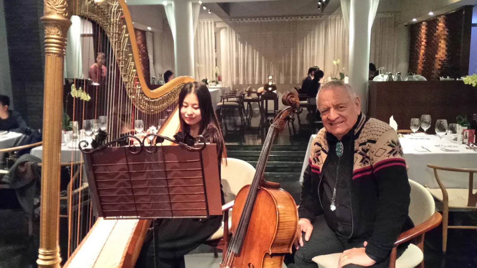 Phil Lane Jr., in a patterned cardigan and turquoise bolo tie, sits smiling beside a young woman playing a golden harp next to a cello in an elegant restaurant.