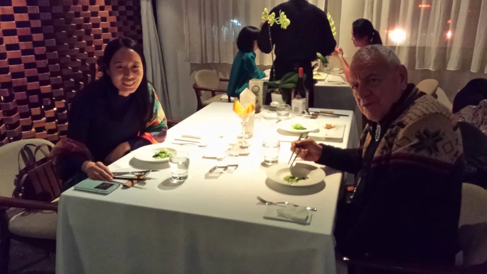 Phil Lane Jr. eats with chopsticks across a white-clothed table from a smiling woman in a dimly lit restaurant with orchids and a carved wooden wall.