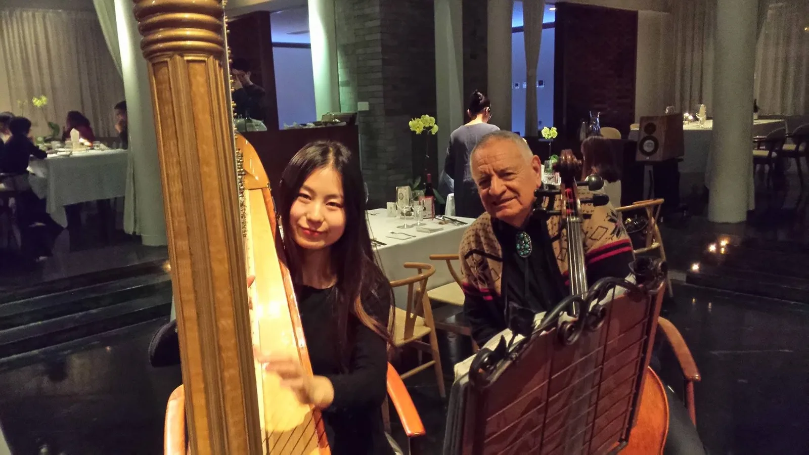 A young harpist smiles beside her harp while Phil Lane Jr. sits next to her behind a cello and music stand in a softly lit restaurant.