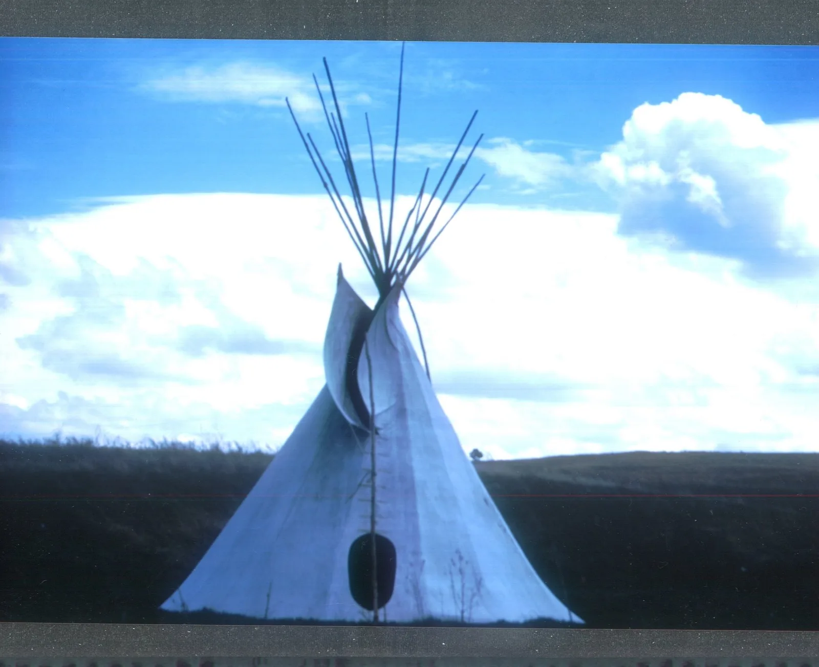 A white canvas tepee with poles crossing at the top and smoke flaps open, standing on dark prairie grassland under a blue sky with large white clouds.