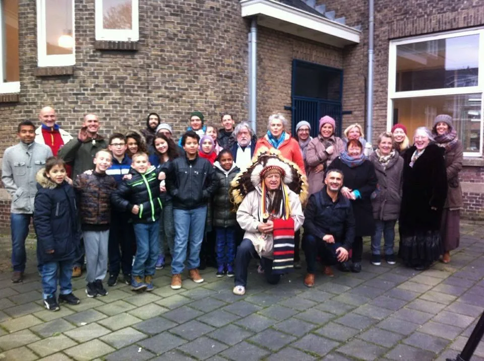 A large group of children and adults in winter coats gathered outside a brick building; an elder in a feathered headdress kneels in front holding a striped cloth.