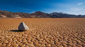 A lone pale rock rests on cracked, dry earth of a desert lakebed, with rugged mountains under a deep blue sky.